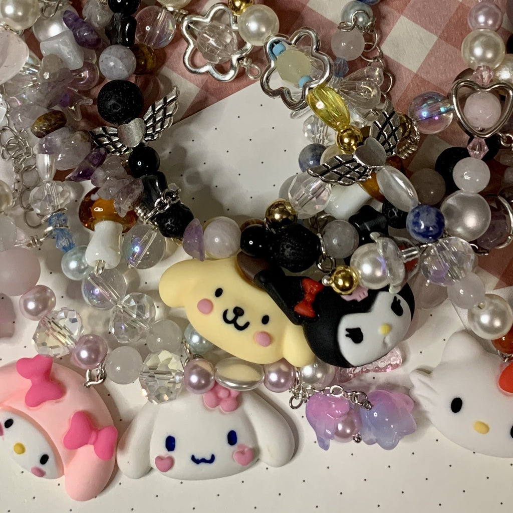 Five Sanrio necklaces lie on top of each other. Characters pictured: Hello Kitty, My Melody, Kuromi, Pompompurin, Cinnamoroll.