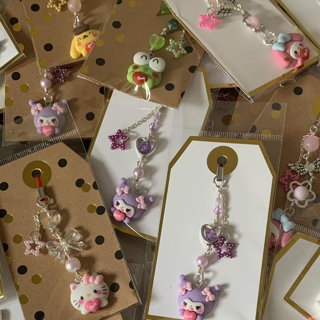 Sanrio phone charms in a brown packaging. Characters pictured: Hello Kitty, My Melody, Kuromi, Pompompurin and Keroppi.